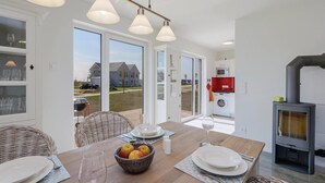 Una cocina moderna con estufa de leña, comedor con mesa para dos personas y vistas a una zona residencial a través de grandes puertas corredizas de vidrio.