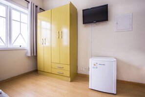 A room with a yellow wardrobe, a white refrigerator, a window with curtains, and a flat-screen TV mounted on the wall.