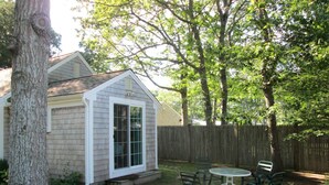Eine kleine Gartenhütte mit Tür und Fenstern, umzäunt mit einem Holzzaun, mit Tisch und Stühlen im Hof.