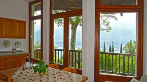 Ein Essbereich mit Tisch für zwei Personen, einer Weinflasche und einem Glas darauf. Zwei Stühle, eine Holzkabine und ein Blick auf eine gebirgige Landschaft durch große Fenster.