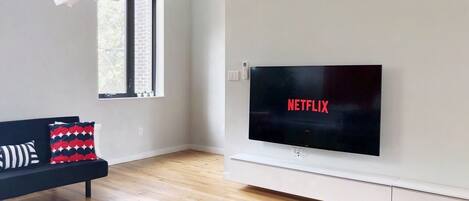 A modern living room with a black sofa, a black rug, a large flat-screen TV, and a window with a view of a brick wall.