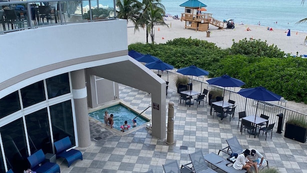 A rooftop pool area with lounge chairs and umbrellas, overlooking a beach with people and a lifeguard stand. - ★★★★★ Hotel 1 Bedroom Suite with ocean view ! Lic :STR-01360 Minimum 3 months.  (Sunny Isles Beach)