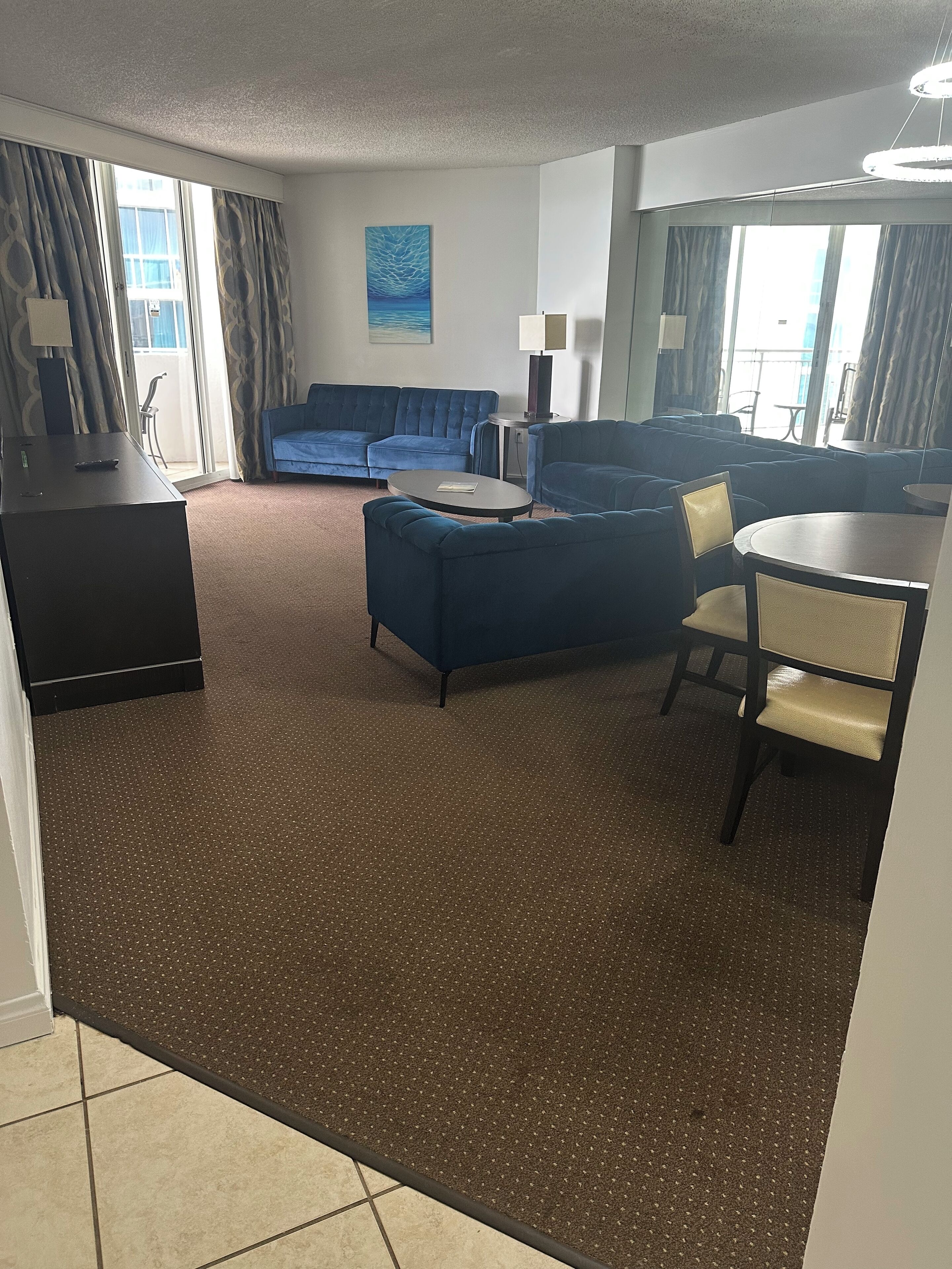 Ein modernes Hotelzimmer mit blauer Sofagarnitur, einem runden Couchtisch, einem Esstisch mit Stühlen und einem großen Fenster mit Vorhängen.