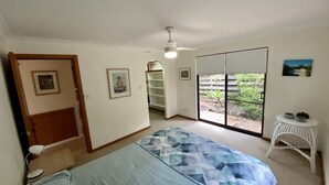 A bedroom with a bed, a ceiling fan, a window with a view of a garden, and a door leading to another room.