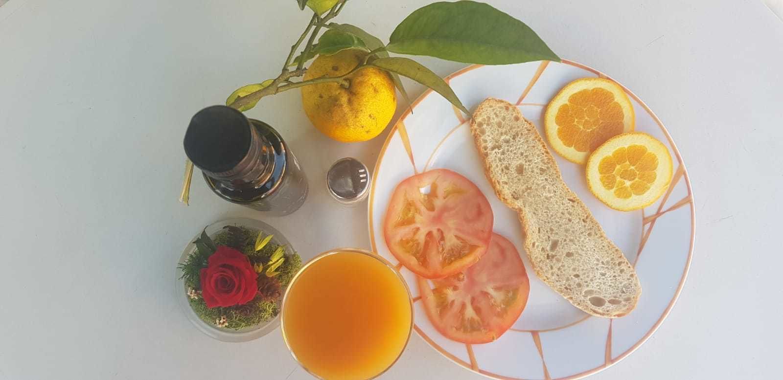 Un plato con rodajas de tomate, gajos de naranja y pan, un vaso de zumo de naranja, un limón y un pequeño adorno con una rosa roja.