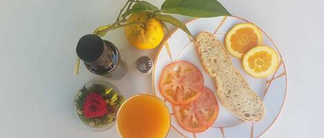 Un plato con rodajas de tomate, gajos de naranja y pan, un vaso de zumo de naranja, un limón y un pequeño adorno con una rosa roja.