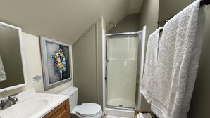 Ein Badezimmer mit einem weißen Waschbecken, einer Toilette und einer Dusche. An der Wand hängt ein gerahmtes Bild und ein weißes Handtuch befindet sich an der Duschkabinentür.