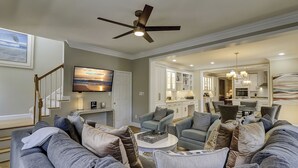 A spacious living room with a large gray sectional sofa, a ceiling fan, and a TV mounted on the wall. - 109 South Forest Beach - Steps away from your toes in the sand! (Hilton Head Island)