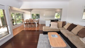A modern living room with a wooden floor, a beige sofa, a dining table with chairs, and a kitchen in the background. - Tranquil Waters - 30 Camden Head Rd Dunbogan (Dunbogan)