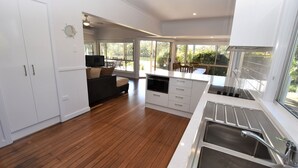 A modern kitchen with a built-in oven, microwave, and sink. There is a dining area with a table and chairs, and a sofa. The room has large windows offering a view of the outdoors. - Tranquil Waters - 30 Camden Head Rd Dunbogan (Dunbogan)