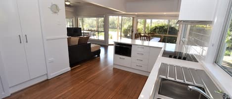 A modern kitchen with a built-in oven, microwave, and sink. There is a dining area with a table and chairs, and a sofa. The room has large windows offering a view of the outdoors.