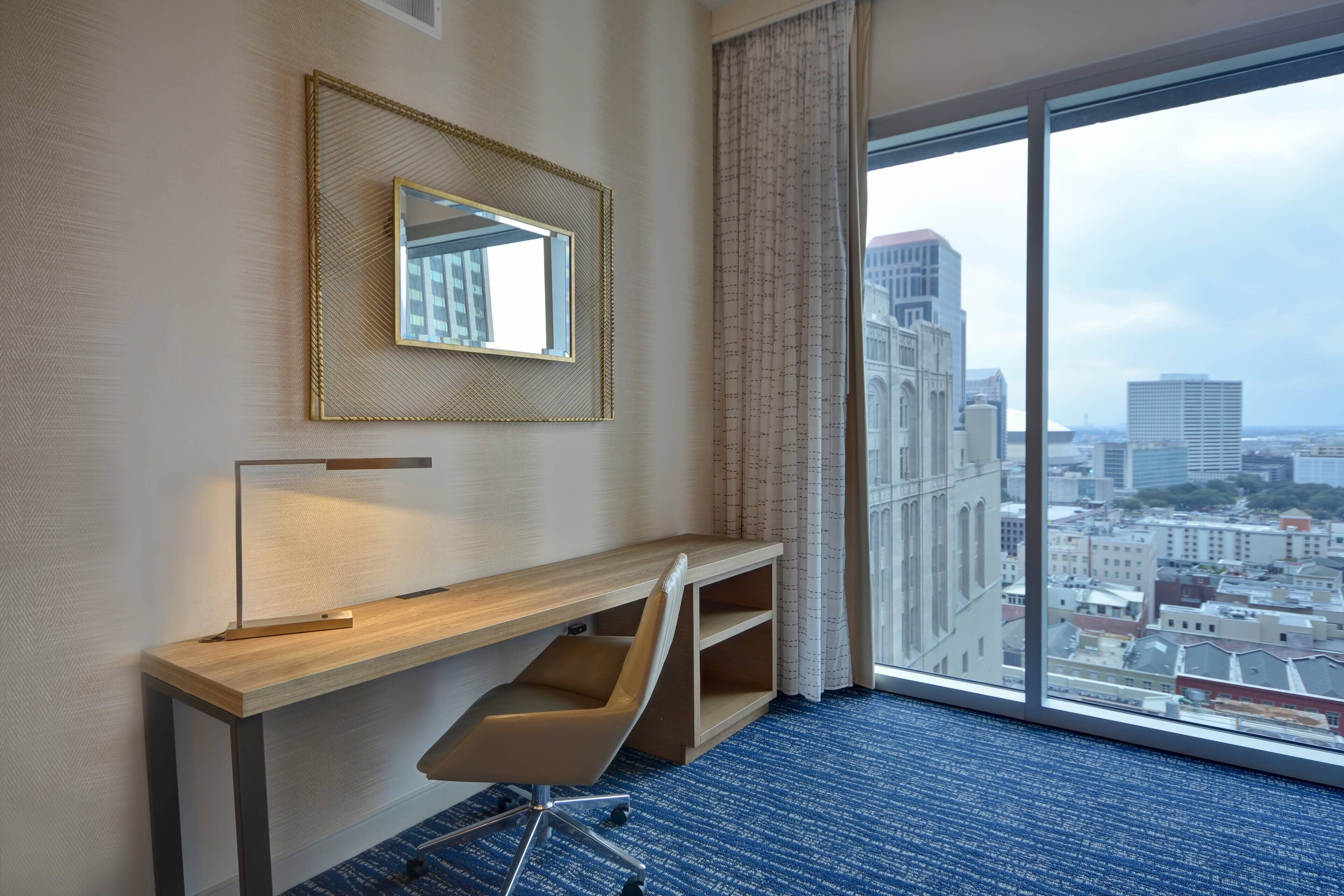 Une chambre d’hôtel dotée d’une grande fenêtre, d’un bureau avec une chaise, d’un miroir et offrant une vue sur le paysage urbain.