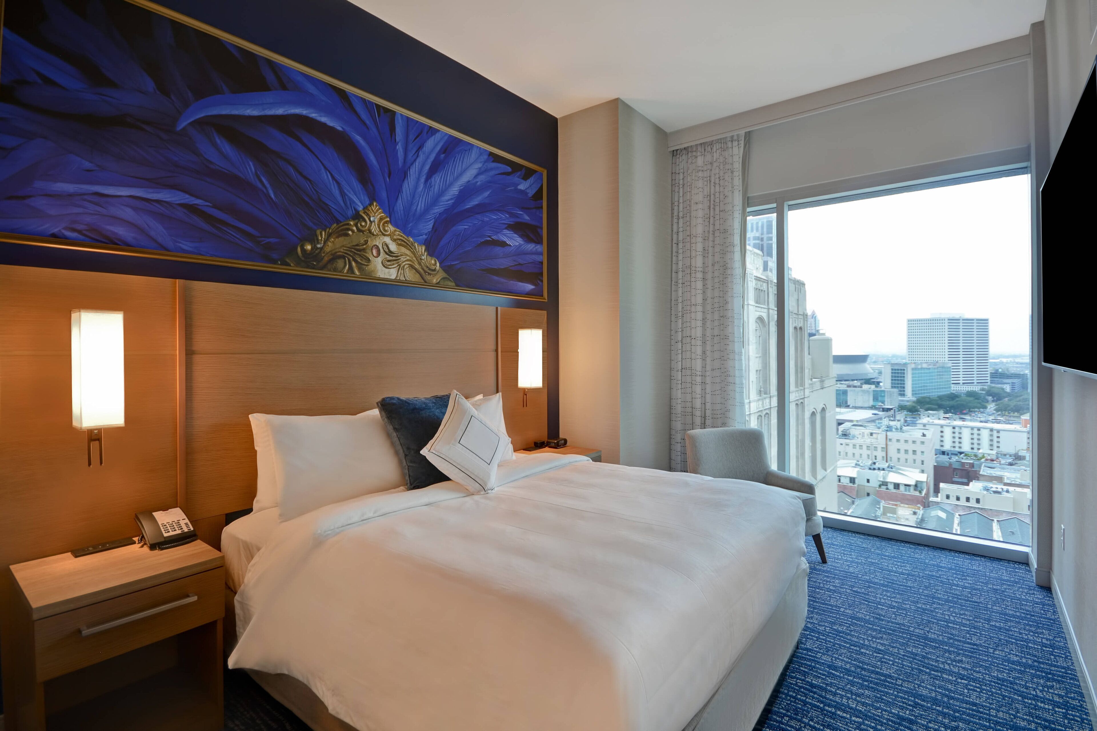 Habitación de hotel con una cama grande, una mesita de noche con teléfono, una silla y un ventanal con vistas a la ciudad.