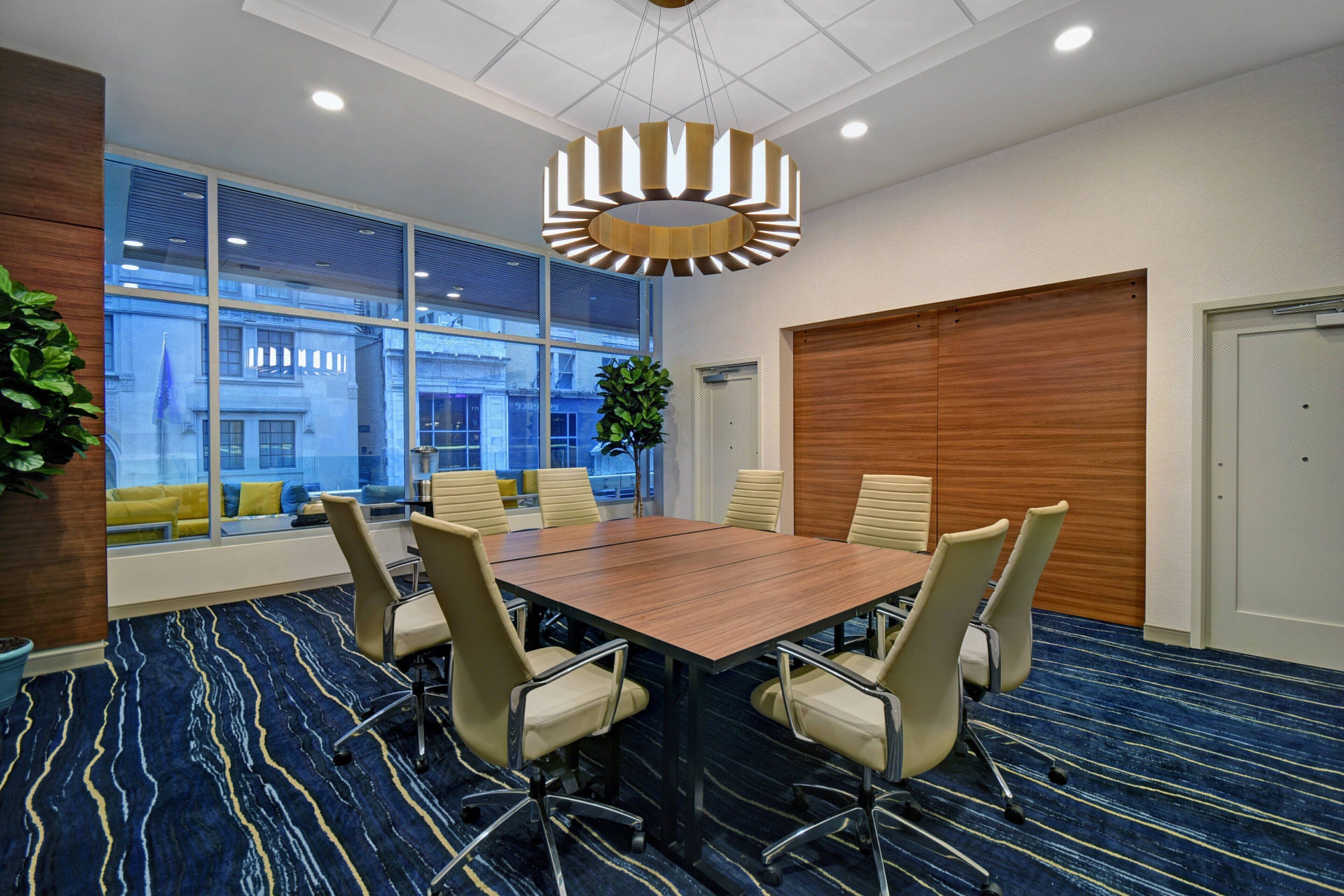 A conference room with a large table, six chairs, a modern chandelier, large windows with a view of buildings, and a patterned carpet.