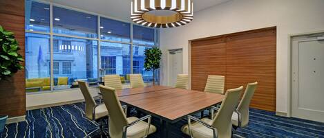 A conference room with a large table, six chairs, a modern chandelier, large windows with a view of buildings, and a patterned carpet.