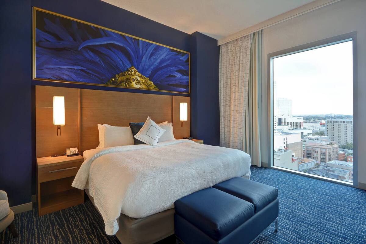 A hotel room with a large bed, a bedside table with a lamp, a window with a city view, and a large painting above the bed.