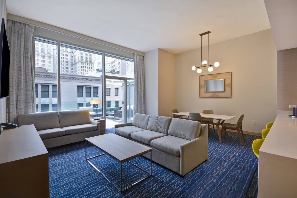 A modern hotel room with a sofa, a coffee table, a dining table with chairs, a TV, and a large window offering a city view.