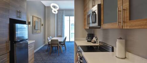 A modern kitchen with a stainless steel refrigerator, microwave, and sink. There is a dining area with a table and chairs, and a blue rug on the floor.