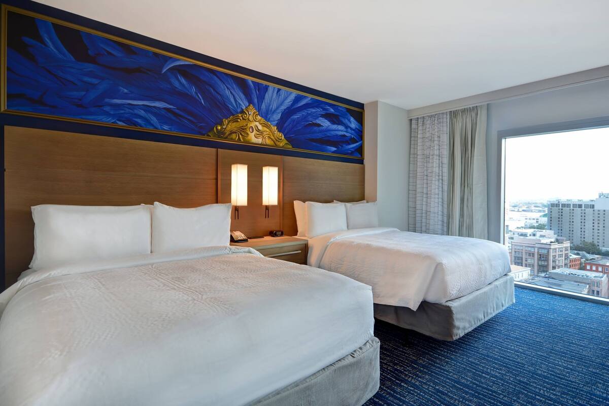 A hotel room with two beds, a large window with a city view, a decorative wall with blue feathers, and a nightstand with a lamp.