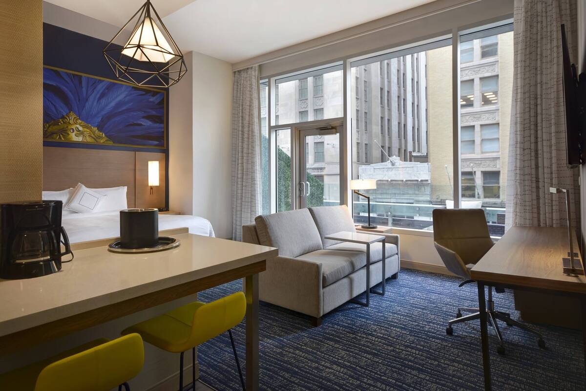 A hotel room with a bed, a sofa, a desk, and a chair. There is a window with a view of buildings outside.