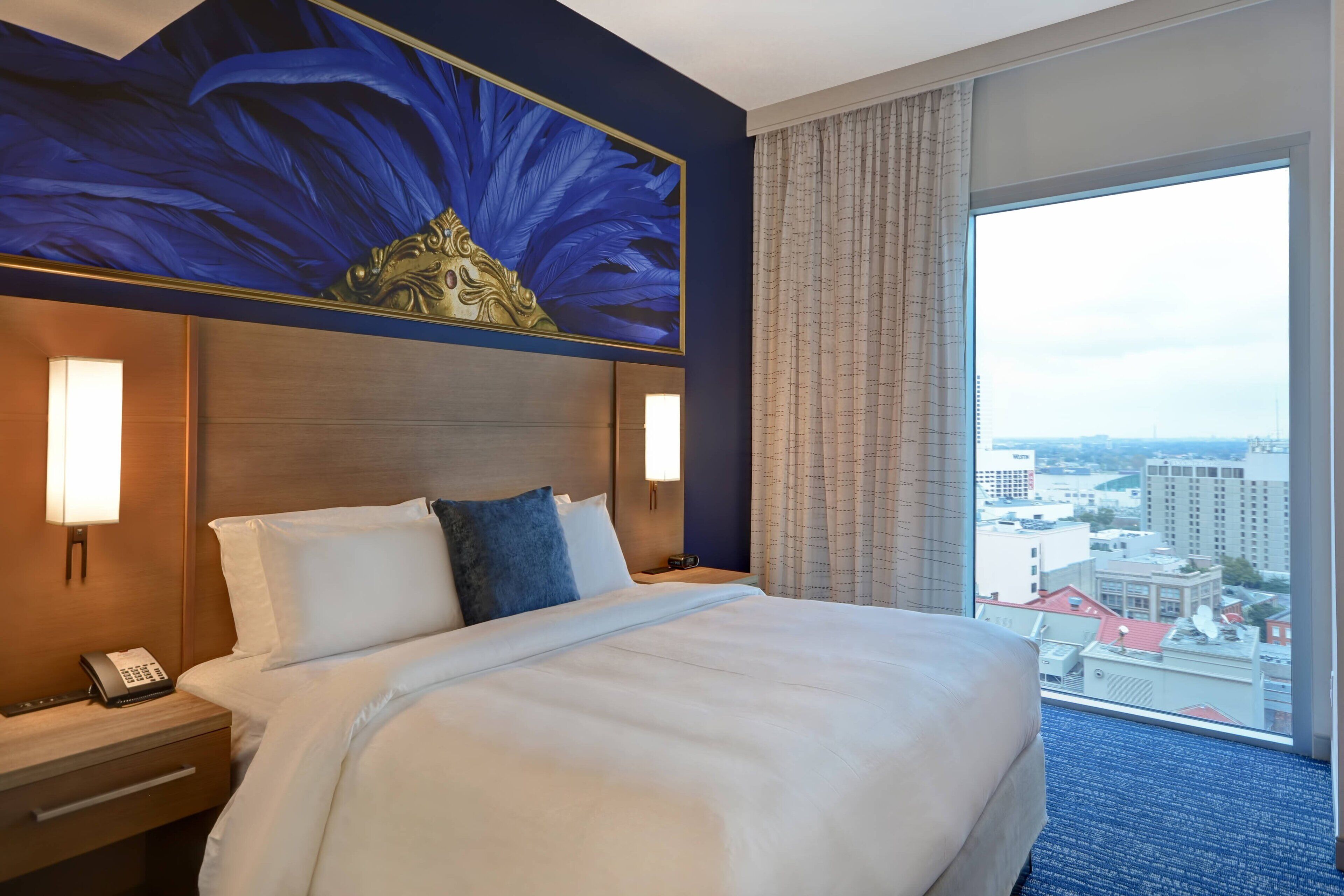 A hotel room with a large bed, a city view, a decorative wall, and a nightstand with a phone.