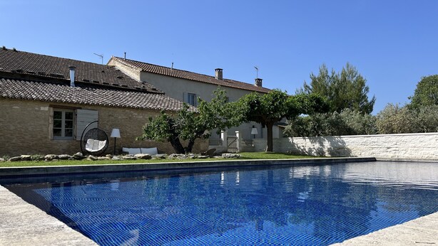 Una piscina con borde de azulejos, un muro de piedra y un edificio con tejado y ventanas aljofadas.