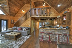 A spacious living room with a large sofa, a wooden coffee table, and a staircase leading to an upper level. A kitchen with a stone island and red bar stools is visible. - Luxury, Toccoa River Front, Fishing, 3 Fireplaces, Kayaks (Mineral Bluff)
