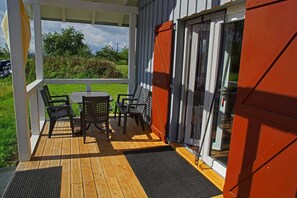A covered outdoor seating area with a table and chairs, a wooden deck, and a glass door leading to a building.