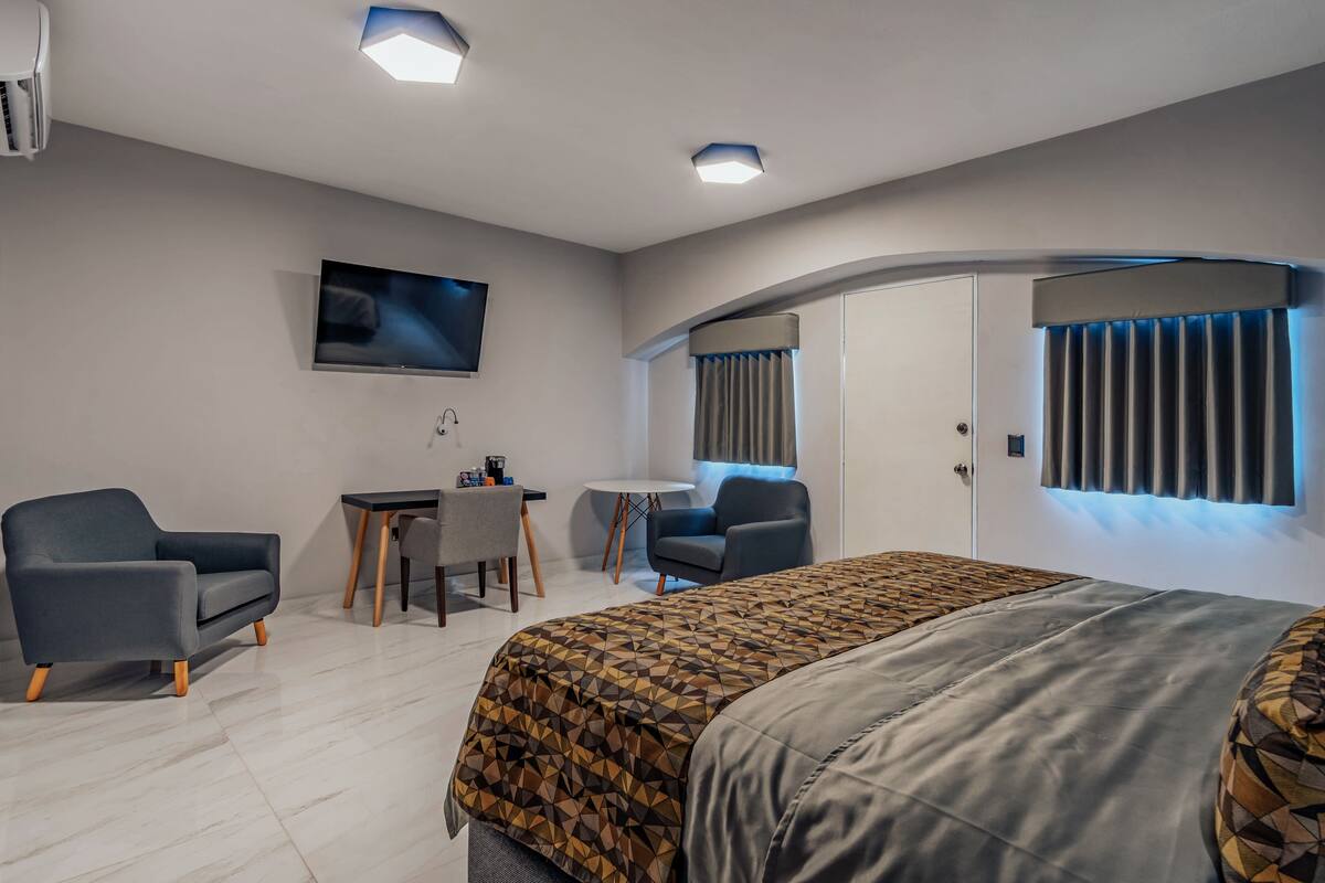 A modern hotel room with a large bed, two armchairs, a small table, a TV, and a curved wall with a window.