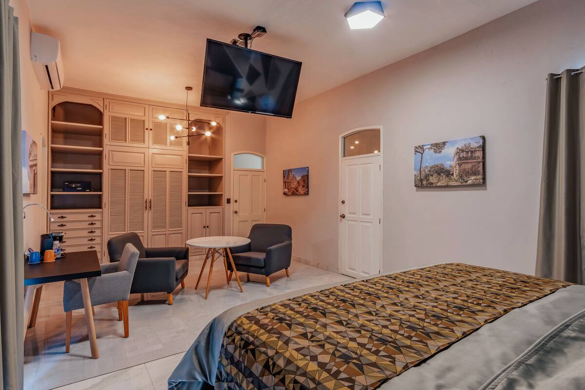 A modern bedroom with a large bed, a TV mounted on the wall, a desk with chairs, and a wardrobe with multiple shelves.