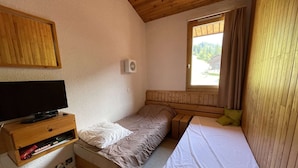 Una habitación pequeña y acogedora con una cama individual, un televisor sobre una estantería de madera y una ventana con cortinas.