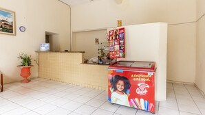 Une salle d’attente avec un sol carrelé, une plante en pot, un téléviseur, un réfrigérateur orné d’une publicité colorée et une horloge murale.