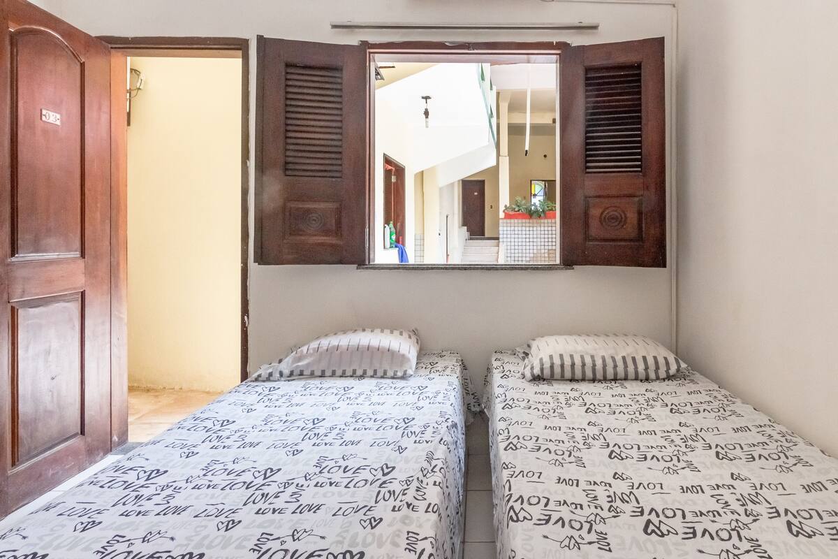 Quarto com duas camas, cada uma com colcha e almofadas estampadas, uma porta de madeira e uma janela com venezianas abertas.