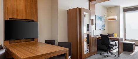 A modern hotel room with a wooden dining table, a flat-screen TV, a large mirror, a desk with a chair, and a large window offering a view.