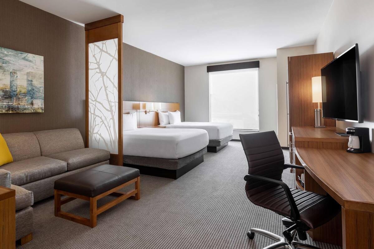 Ein Hotelzimmer mit einer Couch, zwei Betten, einem Schreibtisch mit Fernseher und einem großen Fenster.