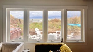 A room with a large window overlooking a beach, a cushioned chair, and a small table. - BEACH Front w/ HOT TUB🔥 Mid Century Home! (Norfolk)