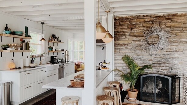 A modern kitchen with white cabinetry, a central island, and bar stools. A stone fireplace with a decorative wreath is visible. - BEACH Front w/ HOT TUB🔥 Mid Century Home! (Norfolk)