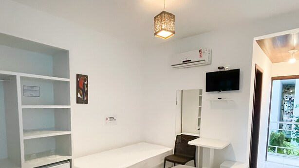 Ein modernes Hotelzimmer mit grauem Sofa, einem kleinen, weißen Kühlschrank, einer eingebauten Sitzbank, einer Badewanne, einem Fernseher und einem großen Fenster mit Blick auf Grünflächen.