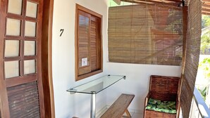 Ein Zimmer mit Holztür, einem Fenster mit Rollläden, einem Glastisch, einer Bank und einem Korbstuhl.