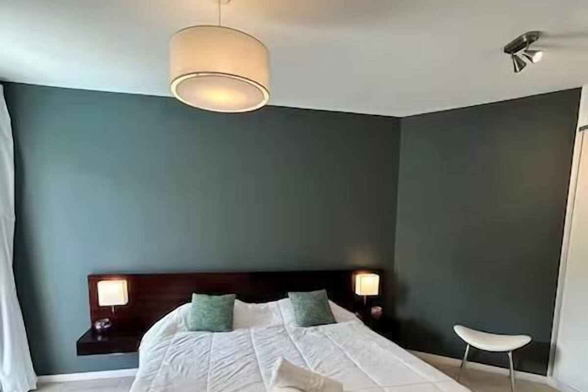 A large bed with a white comforter and pillows, a bedside table with a lamp, and a small white chair.