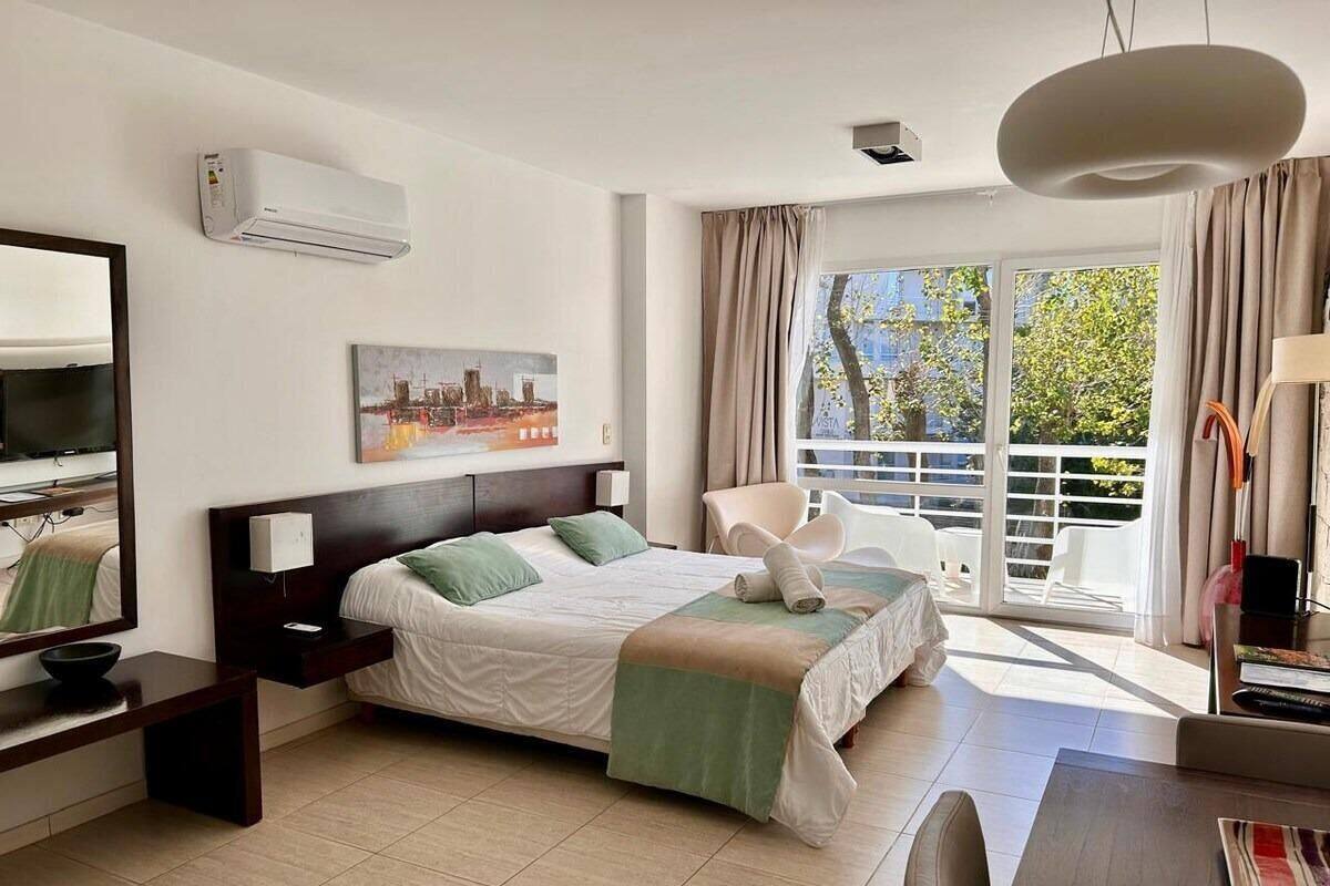 A hotel room with a bed, a desk, a chair, a window with curtains, and an air conditioning unit.
