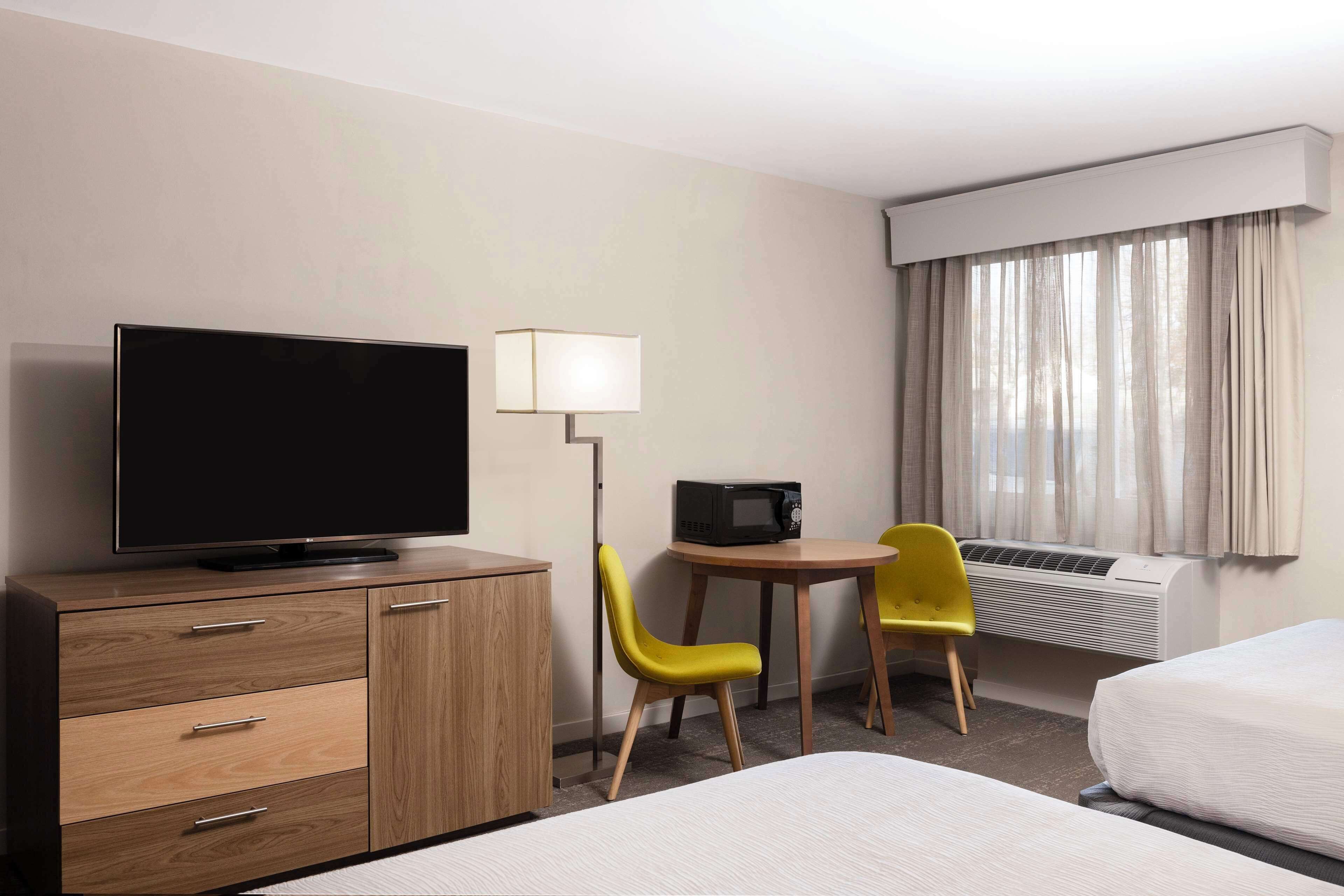 A hotel room with a TV on a wooden cabinet, a small table with a lamp, two yellow chairs, a bed, and an air conditioning unit.