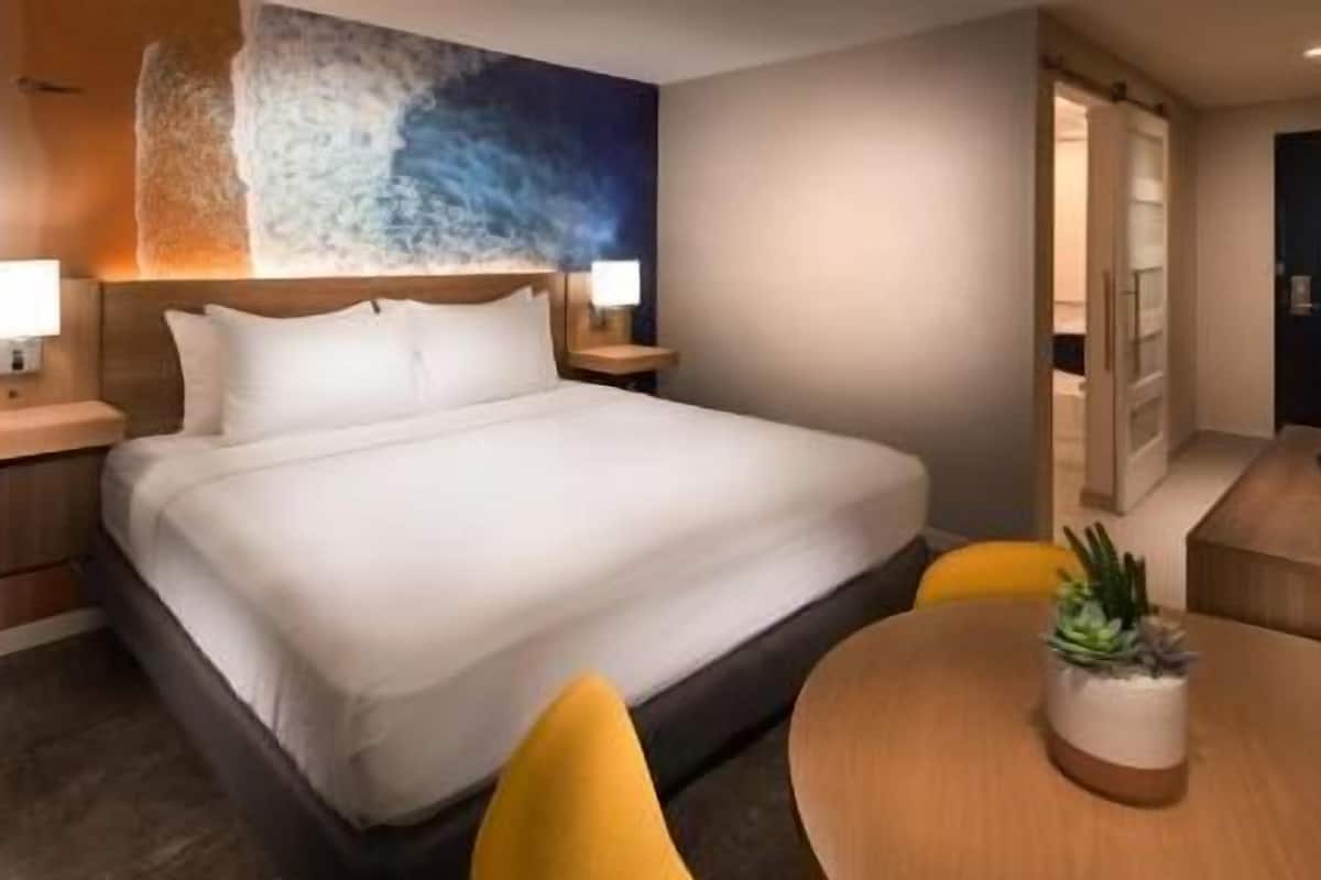 A hotel room with a large bed, bedside tables with lamps, a round table with a potted plant, and a painting on the wall.