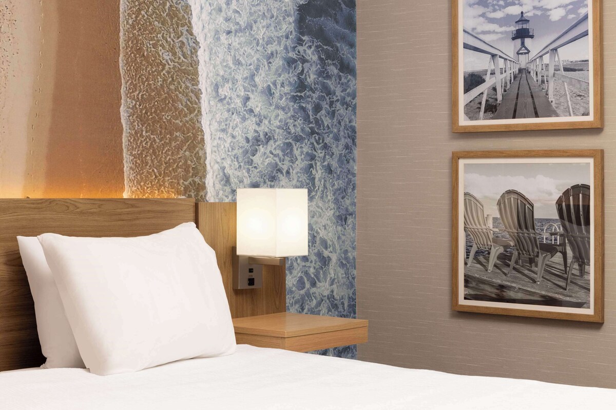 A hotel room with a bed, a nightstand with a lamp, and two framed pictures on the wall.