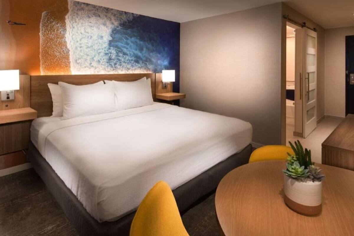 A hotel room with a bed, bedside lamps, a round table with a plant, and a door leading to another room.
