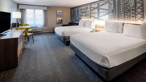 Habitación de hotel con dos camas, un escritorio con televisor, una silla y un tapiz grande en la pared.