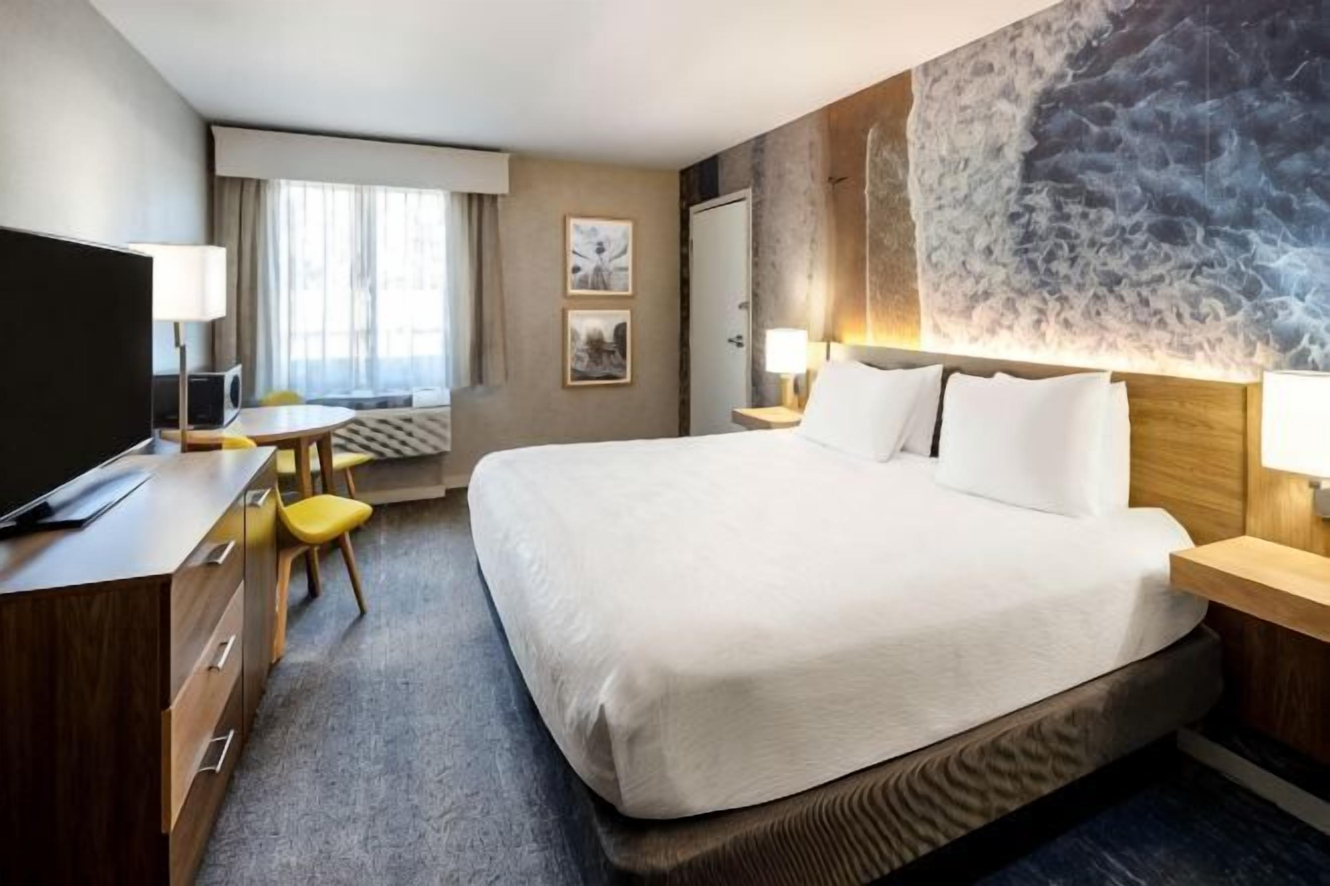 Quarto de hotel com uma cama grande, uma escrivaninha com televisão, uma cadeira, uma luminária de mesa e um quadro na parede.