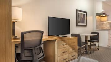 A hotel room with a wooden desk, a flat-screen TV, a chair, a small table, and a lamp.