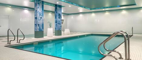 A modern indoor swimming pool with clear blue water, surrounded by a tiled floor and equipped with metal handrails.