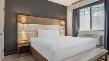 A hotel room with a large bed, bedside lamps, a bench, a window with curtains, and an air conditioning unit.